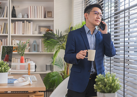 Man in a blazer holding a coffee cup while talking on the phone in a bright office setting with blinds.