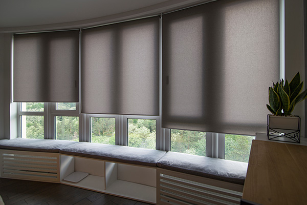 Made-to-measure roller blinds in a spacious window corner with greenery outside.