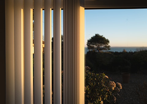 Vertical blinds allowing sunlight in, with a peaceful garden view beyond.