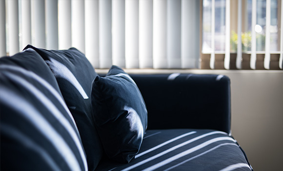Black couch with cushions and vertical blinds casting shadows in a bright room.