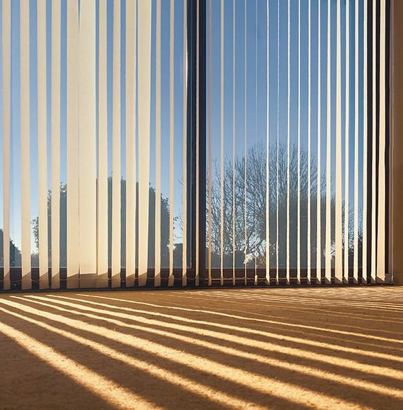 Vertical blinds casting shadows on the floor with a clear sky outside.
