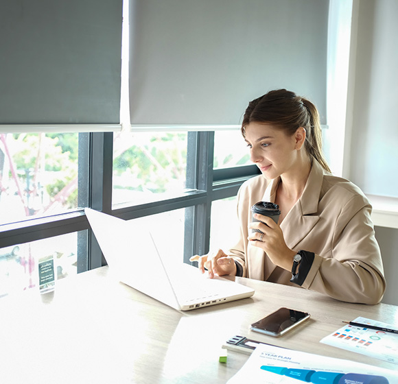 Woman working at desk in bright office with roller blinds and vertical blinds installed on windows