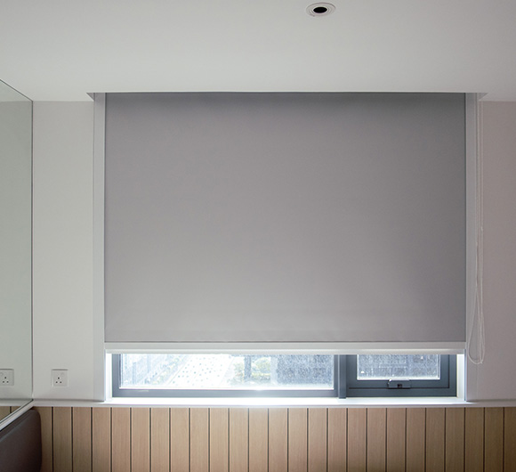 Grey roller blind installed in a window with wooden paneling and clear view outside.