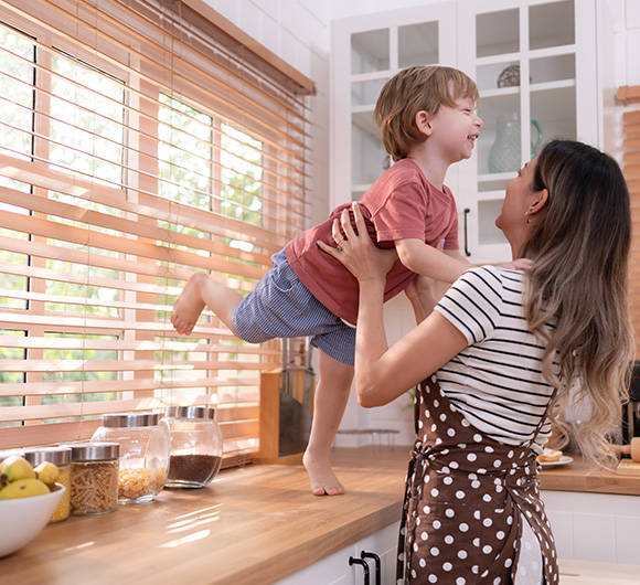 A woman holds a child in front of wooden blinds in a bright kitchen.